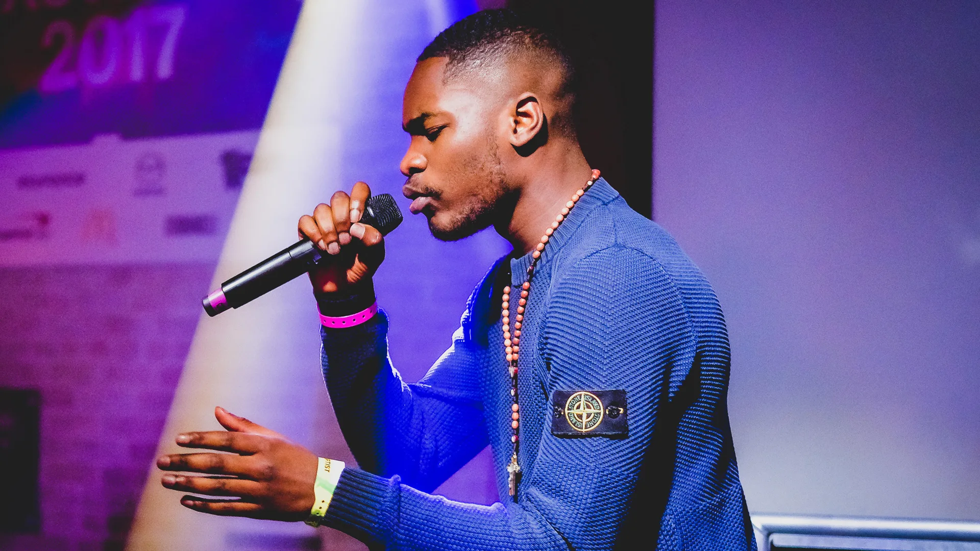 A photo of rapper Dave performing on stage against pink and purple lights. He is wearing a blue top with a neon pink wristband.