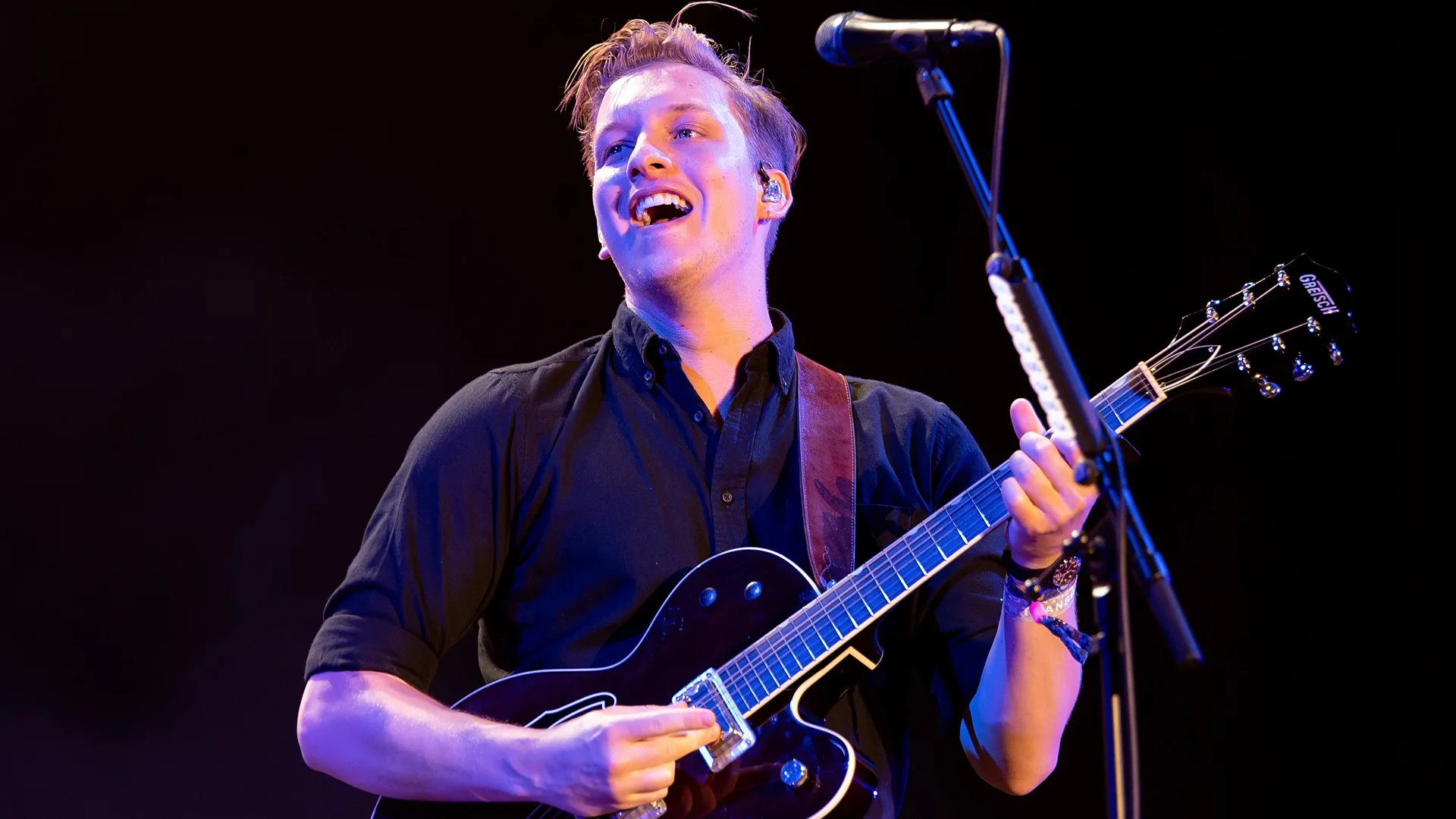 A photo of George Ezra performing on stage with a guitar smiling out to the crowd against a black background.