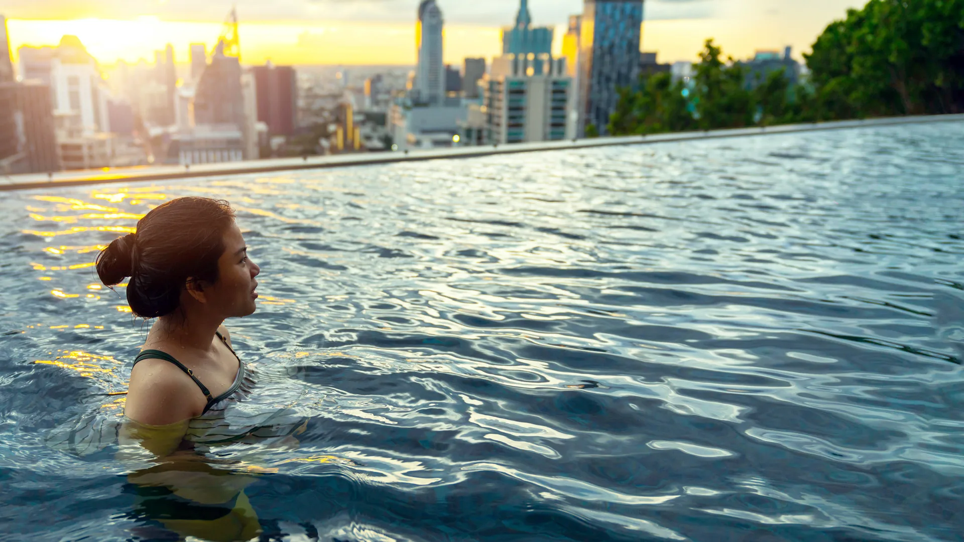 A photo of a woman in a rooftop swimming pool overlooking a city skyline at sunset.