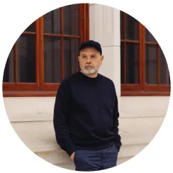 A middle age white man with a cap, trimmed beard and navy sweater  is standing against a wooden window.