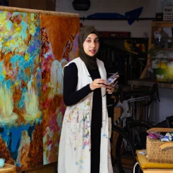 A young woman with a head covering stands in her painting studio.